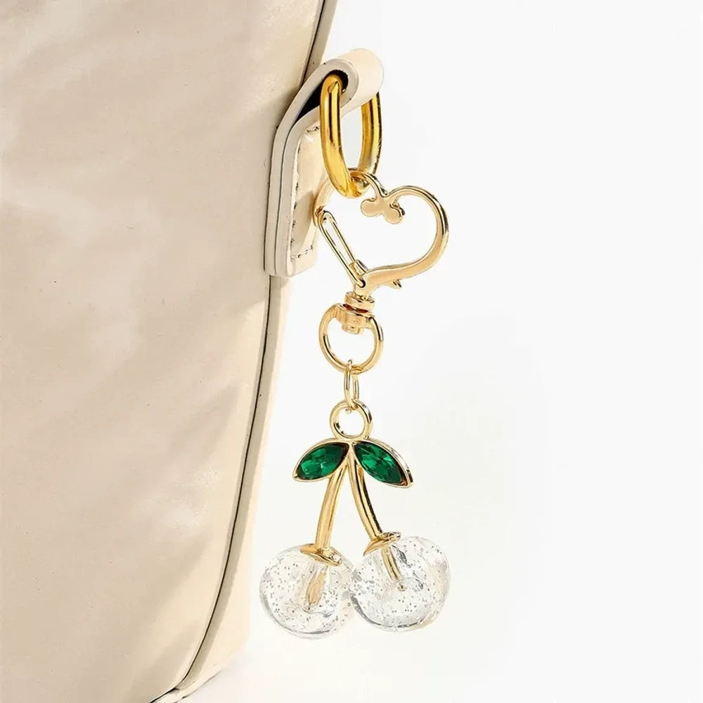 Six crystal glitter cherry charm keychains in red, pink, clear, champagne, purple, and blue with gold heart-shaped lobster clasps and green enamel leaves on white background. - Clear