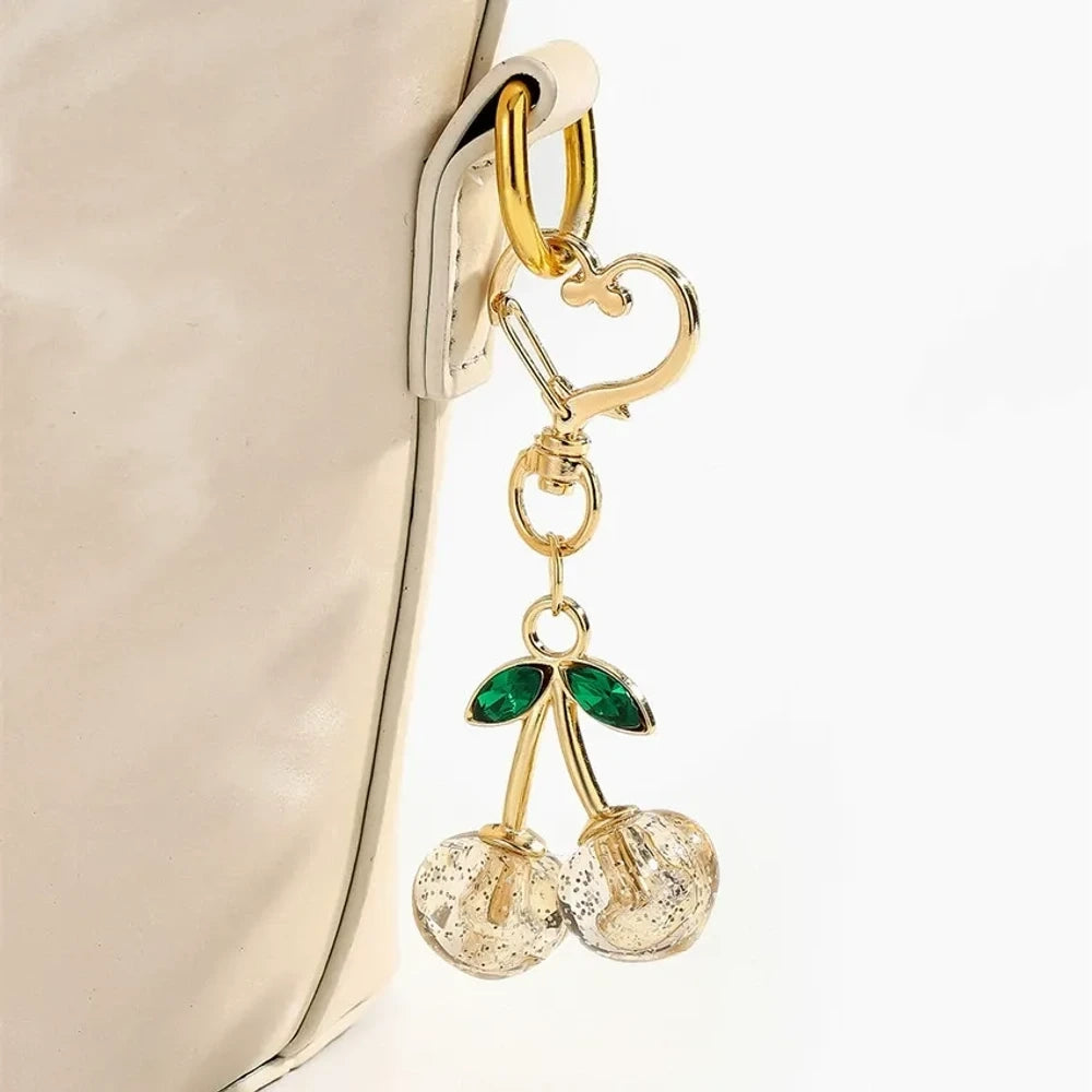 Six crystal glitter cherry charm keychains in red, pink, clear, champagne, purple, and blue with gold heart-shaped lobster clasps and green enamel leaves on white background. - Gold