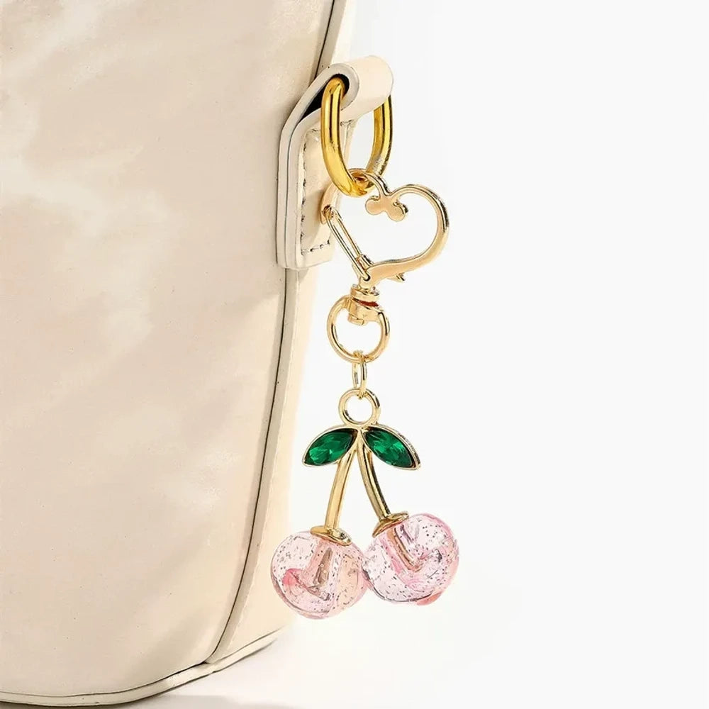 Six crystal glitter cherry charm keychains in red, pink, clear, champagne, purple, and blue with gold heart-shaped lobster clasps and green enamel leaves on white background. - Pink