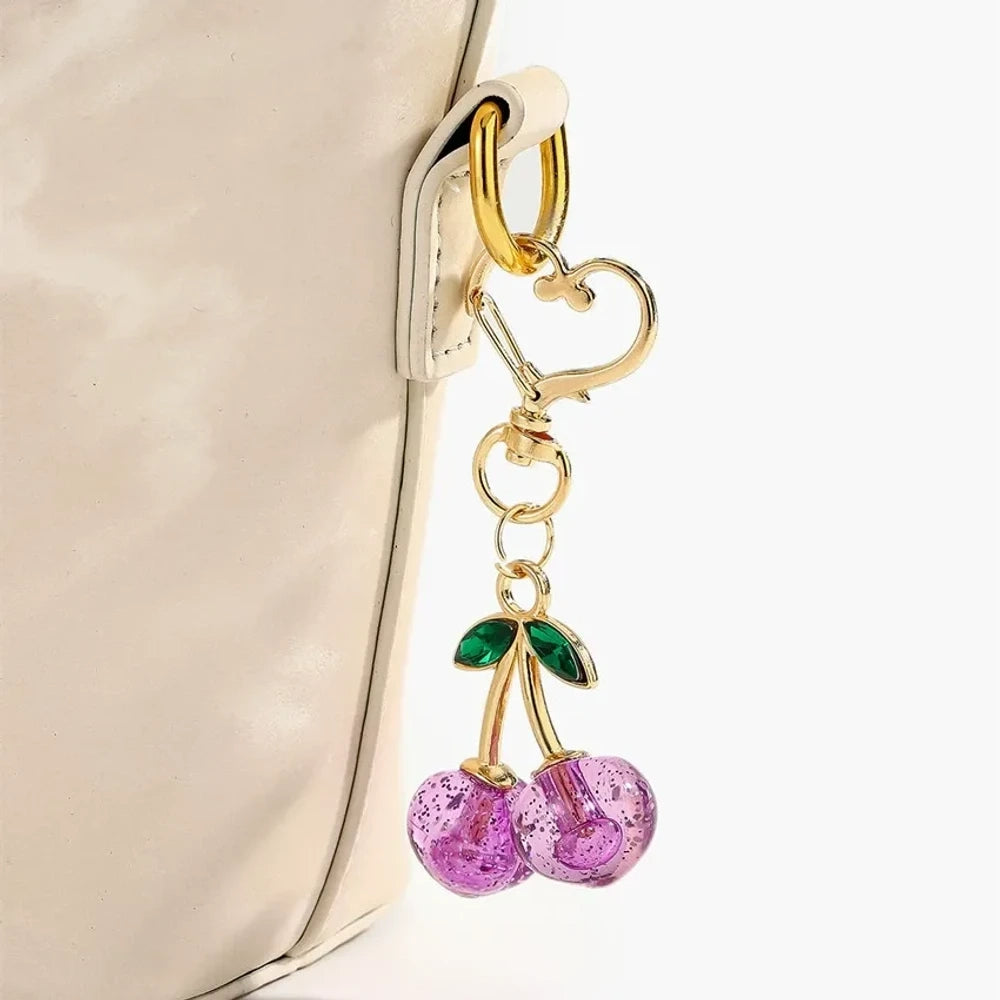 Six crystal glitter cherry charm keychains in red, pink, clear, champagne, purple, and blue with gold heart-shaped lobster clasps and green enamel leaves on white background. - Purple