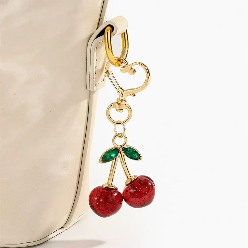 Six crystal glitter cherry charm keychains in red, pink, clear, champagne, purple, and blue with gold heart-shaped lobster clasps and green enamel leaves on white background. - Red