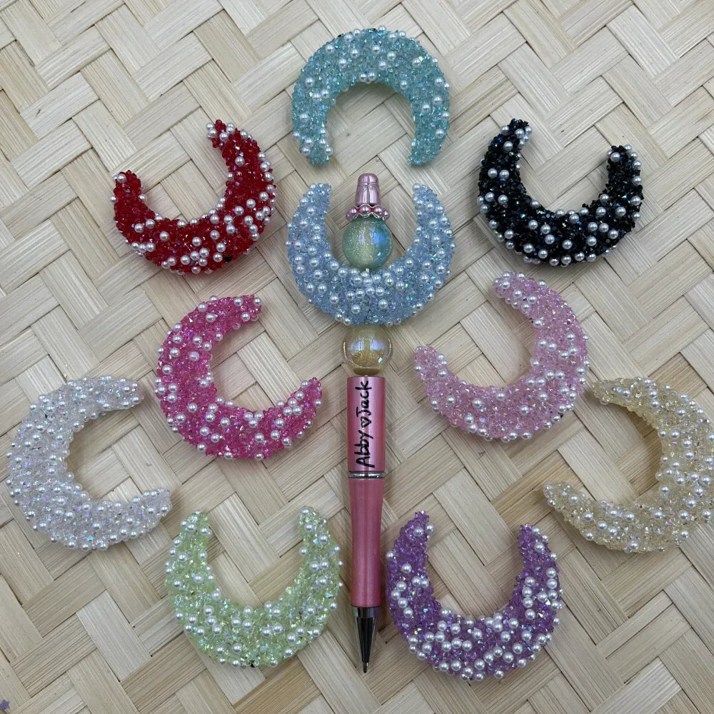 Assorted crystal and pearl crescent moon shaped fancy beads in red, blue, black, pink, white, green, purple, and cream arranged around a pink beadable pen on wicker background.