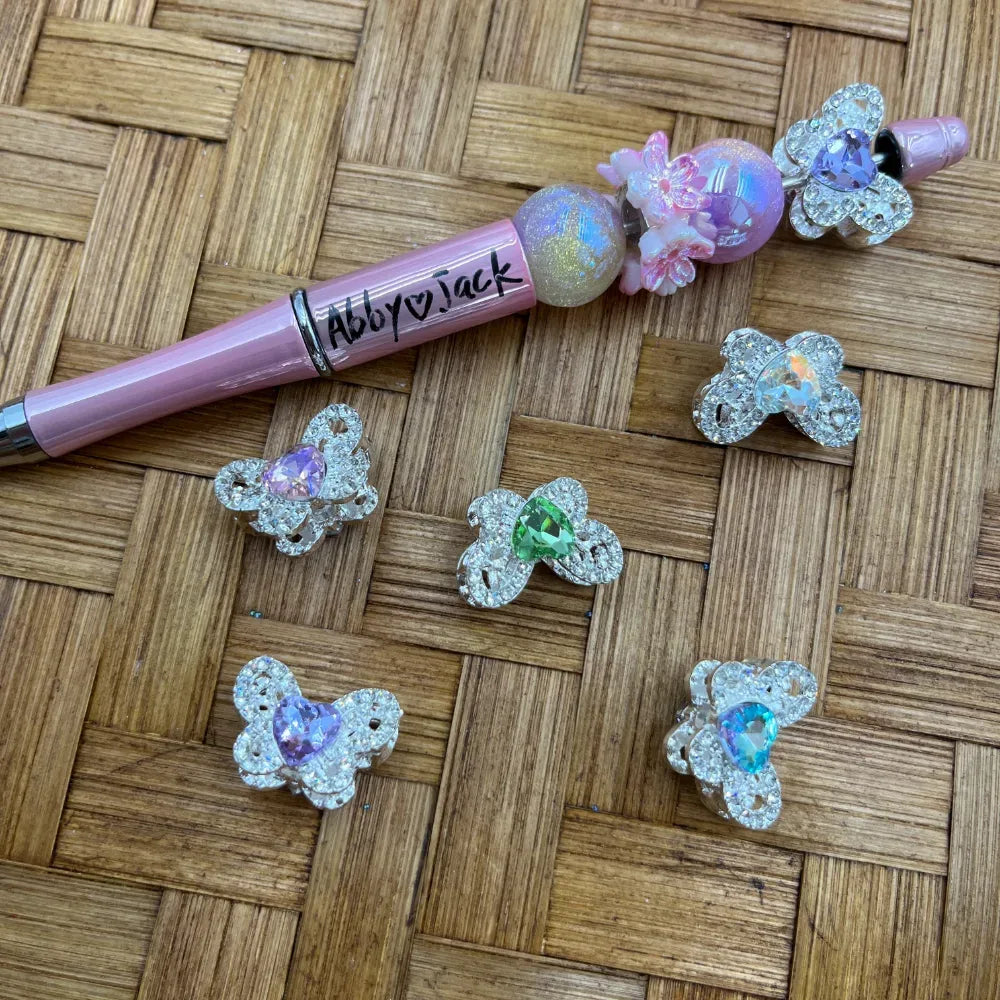 Silver crystal rhinestone butterfly bear shaped metal beads with colorful teardrop gemstone centers in purple, green, blue, and aurora borealis, displayed on woven bamboo surface next to a decorated pink beadable pen labeled Abby and Jack.