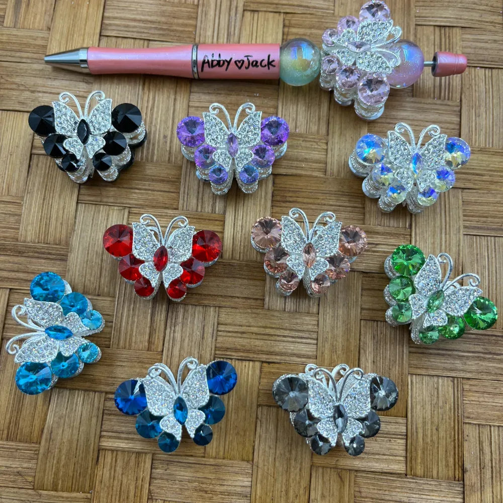 Assorted crystal rhinestone butterfly fancy beads in multiple colors including black, purple, red, pink, aqua, blue, gray, green, and iridescent AB, displayed on a woven surface next to a beadable pen for size reference.