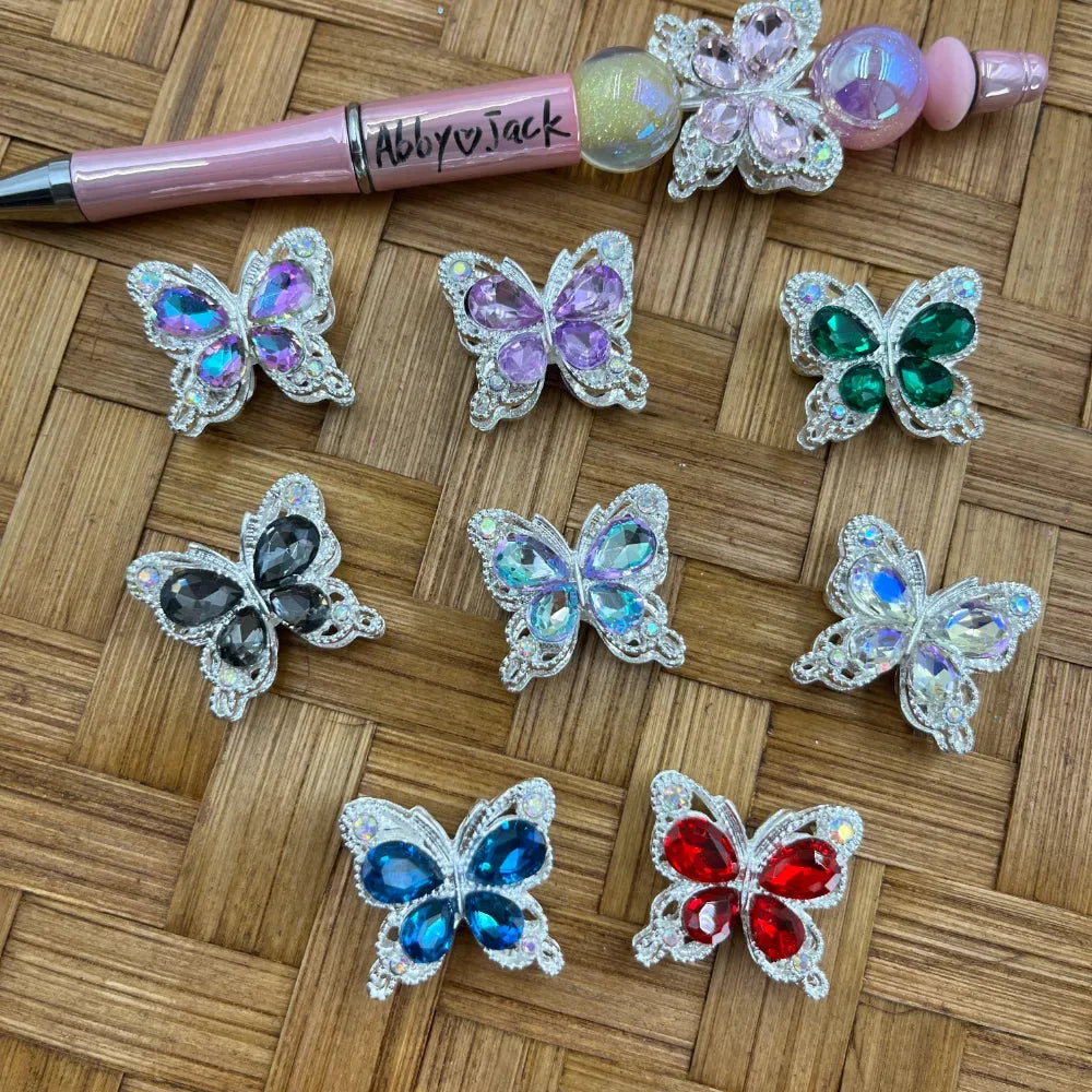 Eight crystal rhinestone butterfly metal beads in assorted colors including purple, lavender, green, black, aqua, iridescent, blue, and red, displayed on woven surface next to a pink beadable pen.