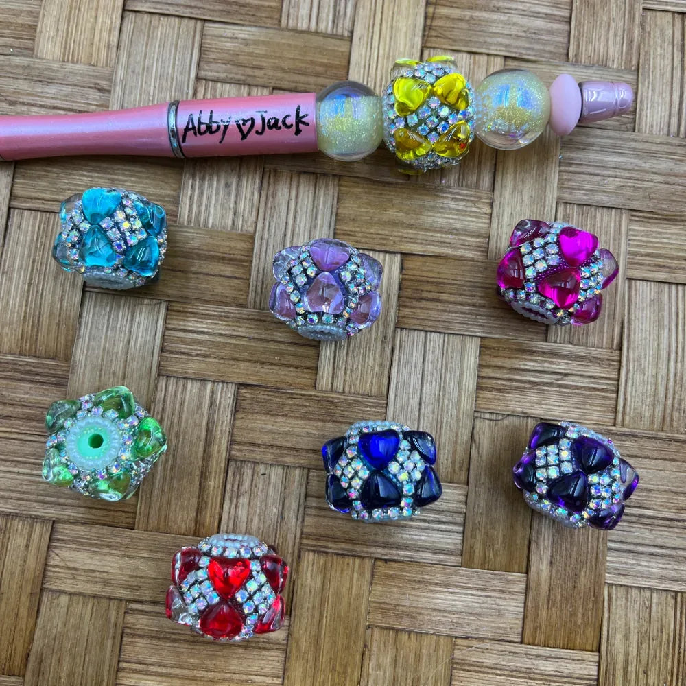Assorted crystal rhinestone flower cluster fancy beads in teal, lavender, yellow, pink, green, red, blue, navy, and purple displayed on woven bamboo surface next to a branded Abby Jack beadable pen.