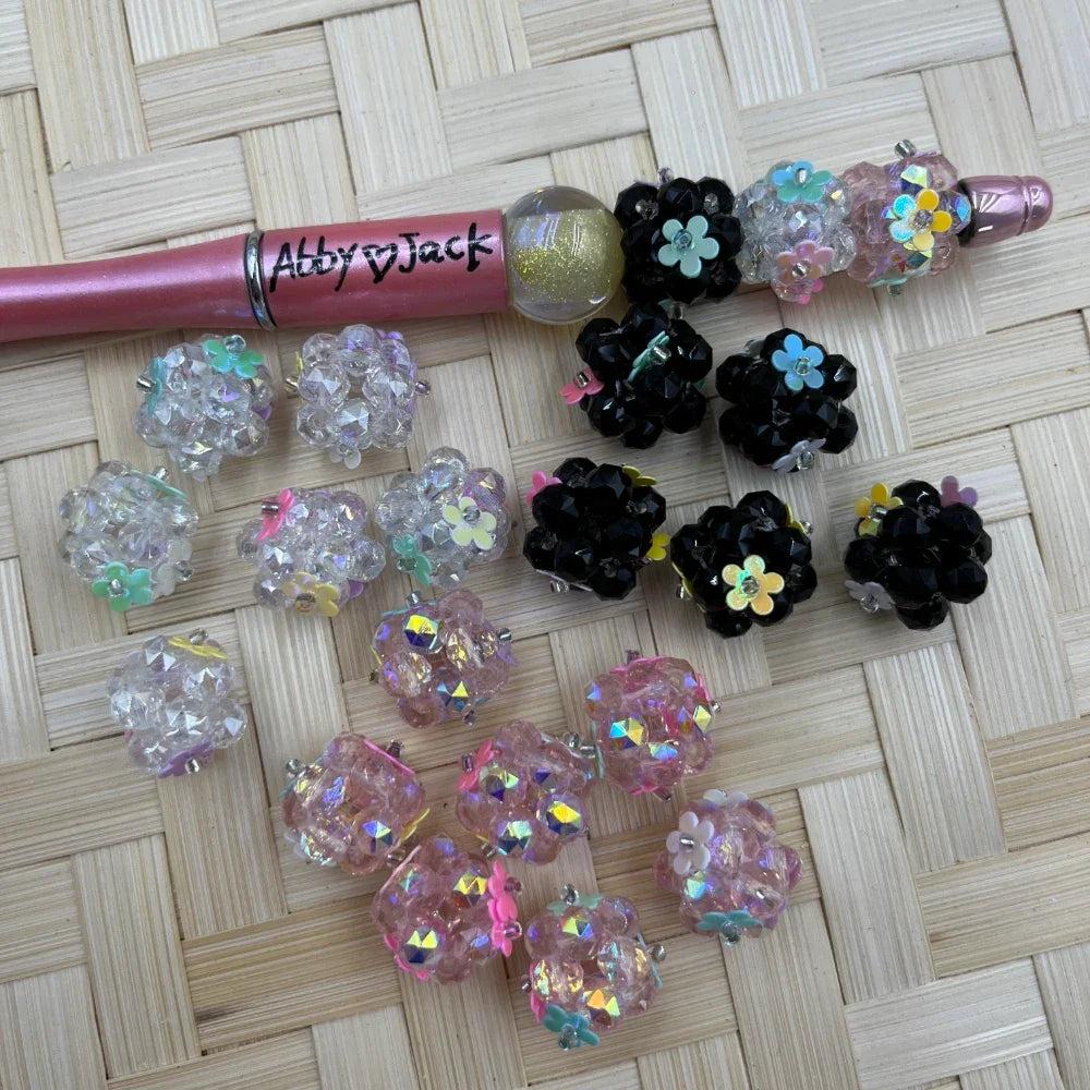 Assorted crystal rhinestone flower cluster fancy beads in clear, black, and pink with iridescent floral accents displayed on woven surface beside a pink beadable pen labeled Abby Jack