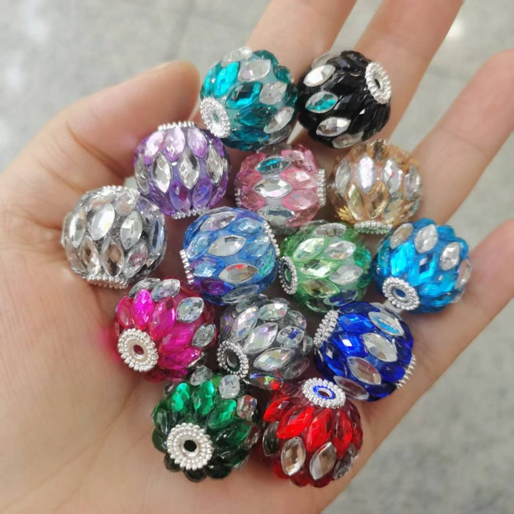 Hand holding a colorful assortment of round crystal rhinestone fancy beads in multiple colors including teal, purple, pink, blue, green, red, black, and gold with silver accents and marquise-cut rhinestone embellishments.