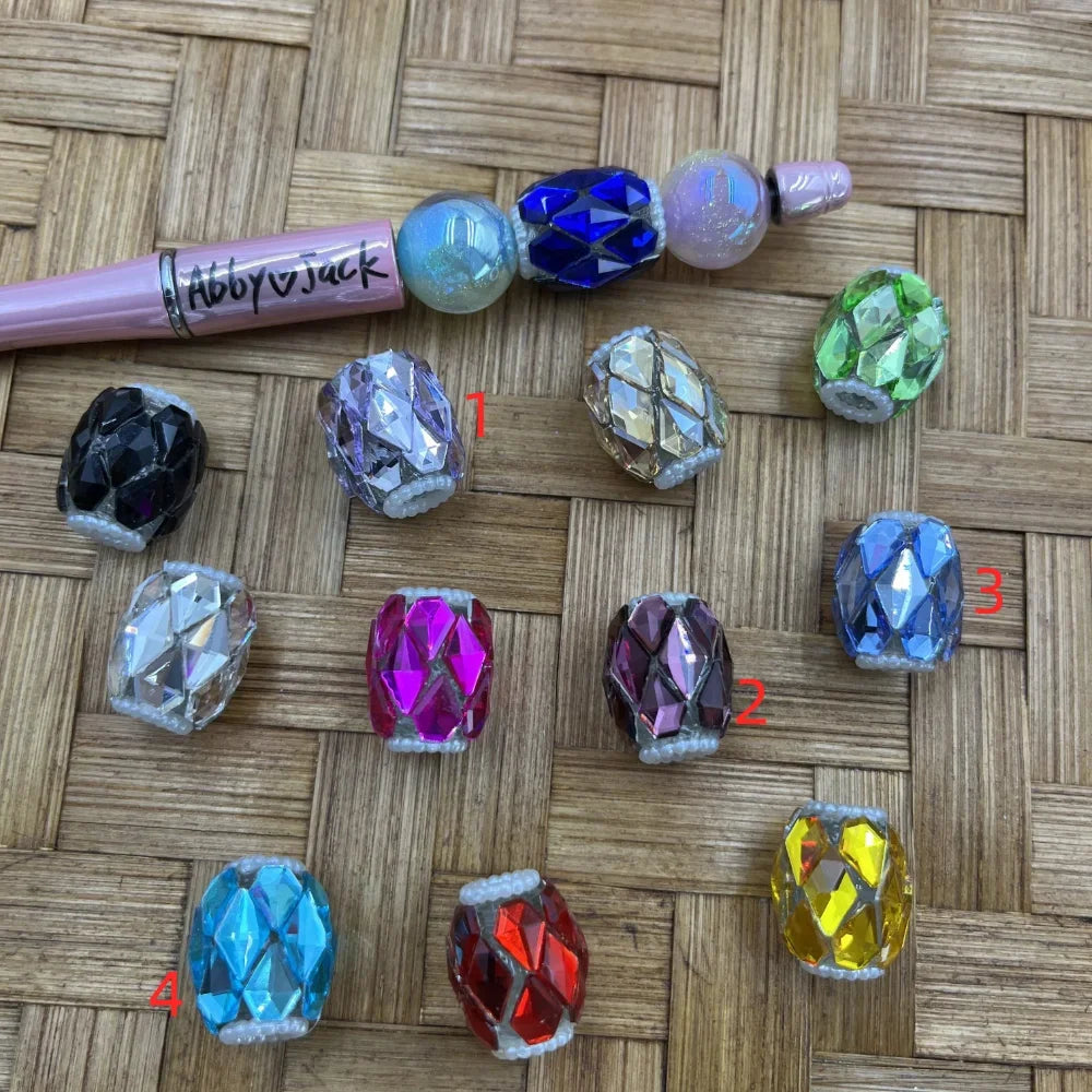 Assorted crystal rhinestone mesh barrel fancy beads in 12 colors including black, clear, purple, gold, green, pink, magenta, blue, red, yellow, and iridescent, displayed on a woven surface next to a pink beadable pen from Abby and Jack brand.