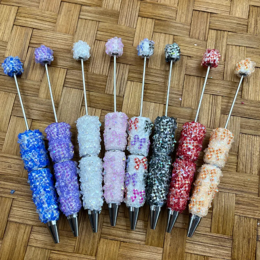 Eight colorful crystal rhinestone sugar beadable pens in blue, purple, white, pink, mixed, black, red, and orange displayed on woven bamboo background at DIY Mini Planet.