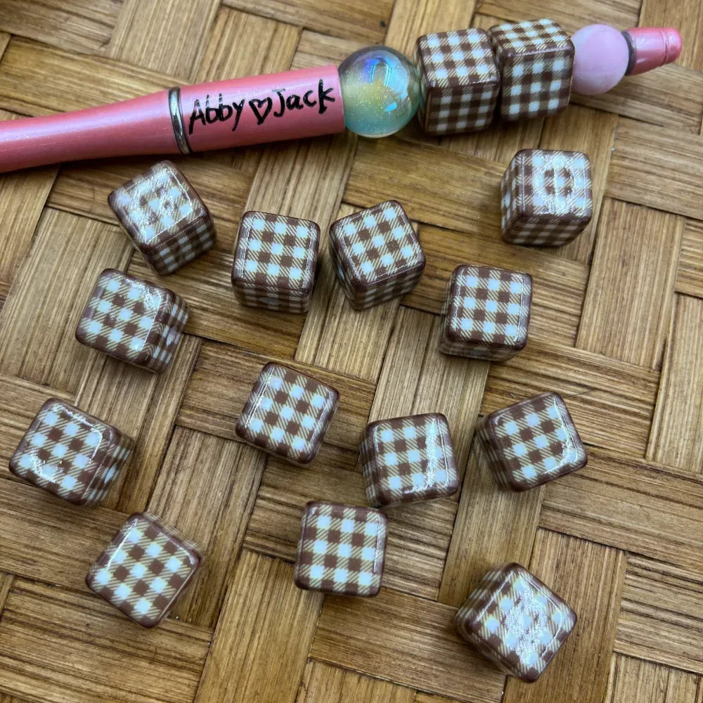 Brown and white gingham plaid patterned cube acrylic beads displayed on a woven surface next to a pink beadable pen, showing checkered pattern detail on all sides.