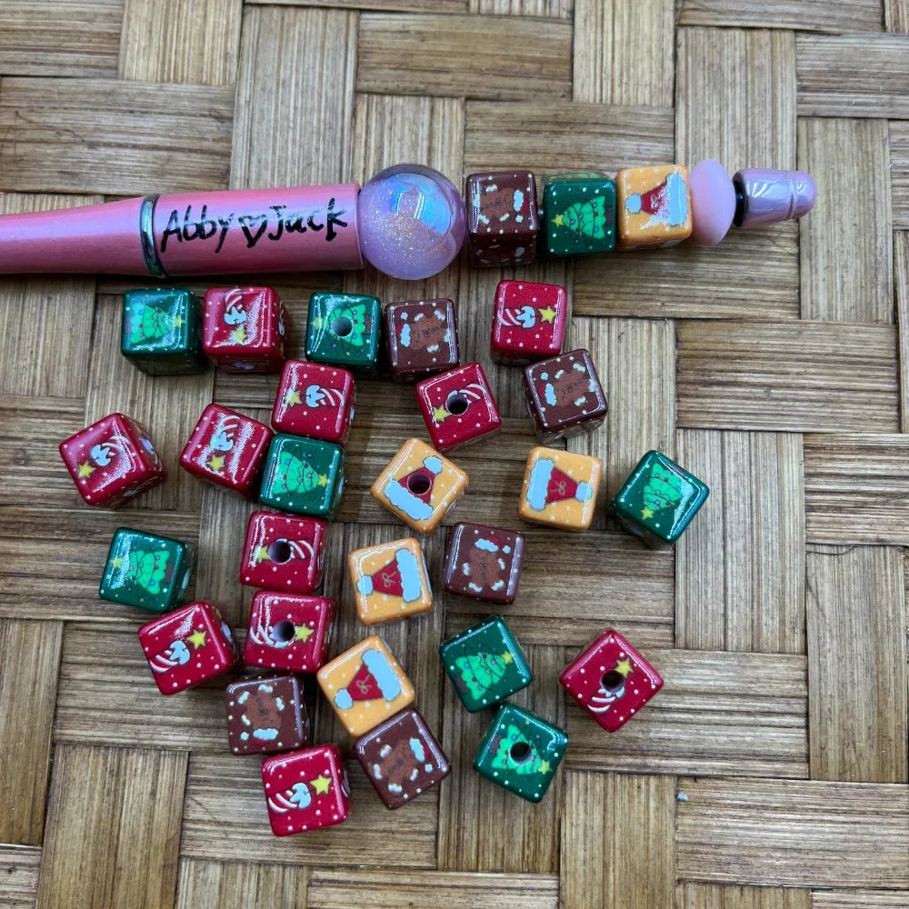 Colorful Christmas-themed cube acrylic beads in red, green, brown, and orange with Santa hat, Christmas tree, and holiday designs, shown with a pink beadable pen on a wicker surface.