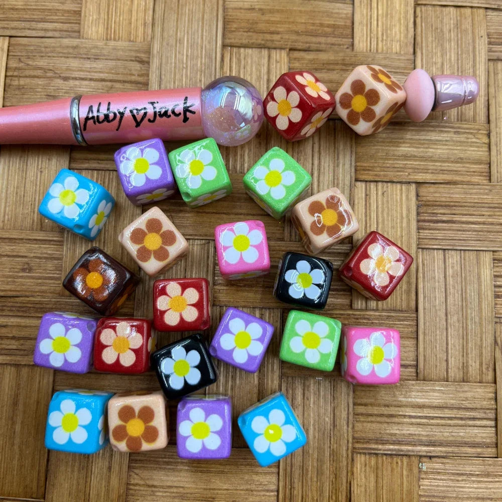 Colorful cube acrylic beads with white daisy flower print in mixed colors including blue, purple, pink, red, black, green, and peach, shown next to a beadable pen on a wicker surface.