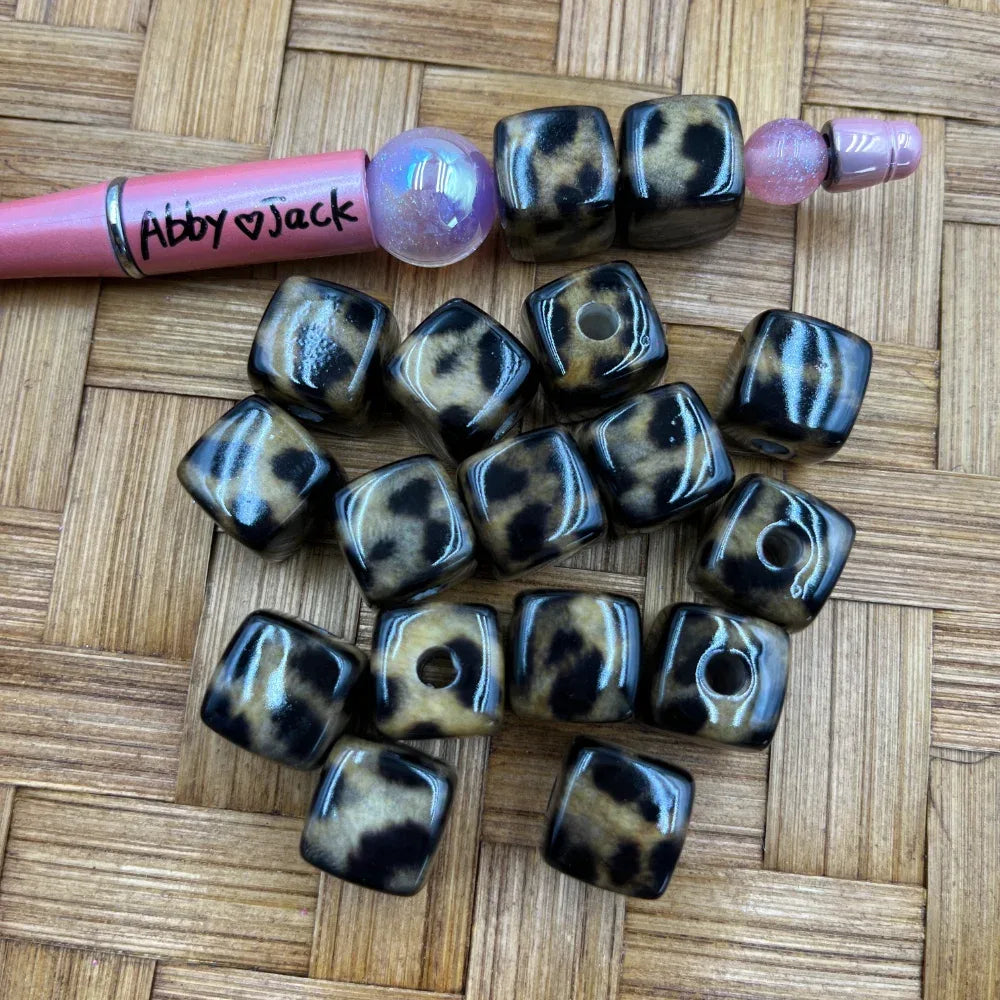 Glossy black and tan leopard print cube-shaped acrylic beads scattered on a woven surface, shown next to a beadable pen for size reference.