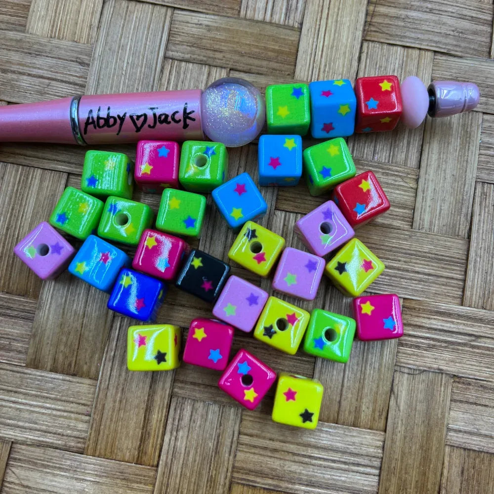 Assorted colorful cube acrylic beads with multicolor star prints in neon green, pink, yellow, blue, black, and purple, shown with a pink beadable pen on a wicker surface.
