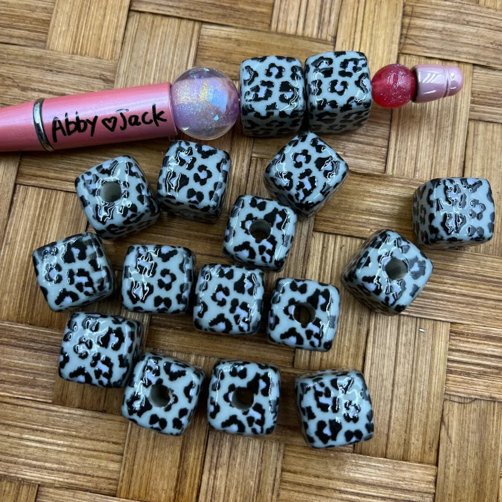 White and black leopard print cube acrylic beads with center hole, shown next to beadable pens on a wicker surface, perfect for DIY jewelry making and beadable pens.