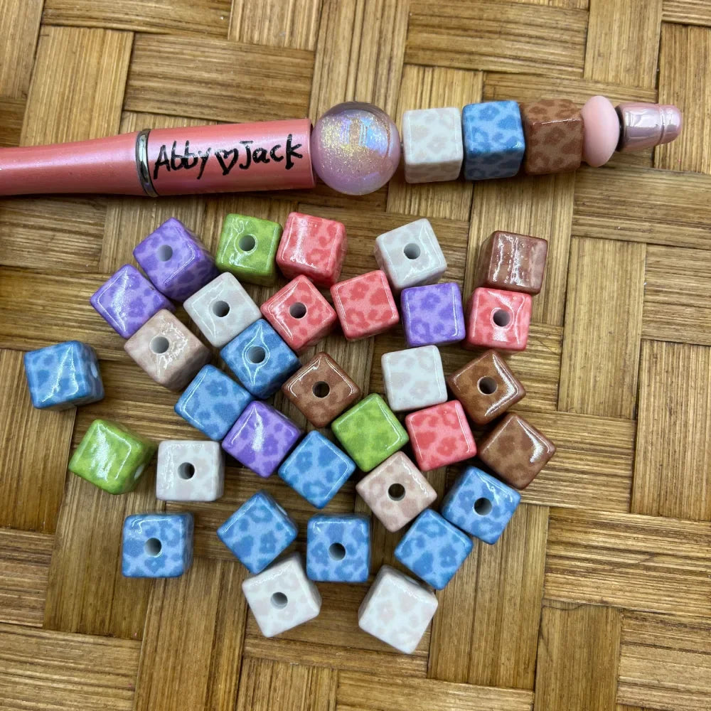 Colorful cube square leopard print acrylic beads in pastel blue, purple, pink, green, brown, and white scattered on a wicker surface next to a pink beadable pen labeled Abby Jack.