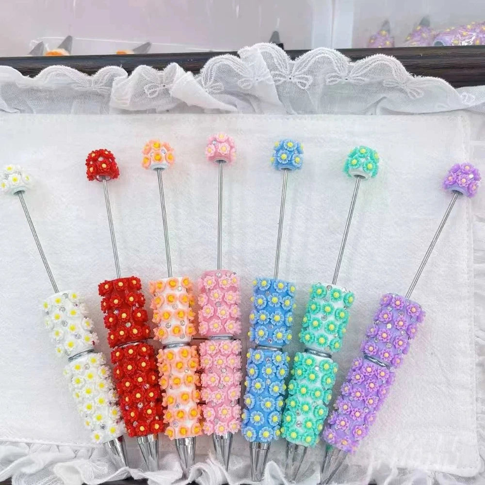 Seven colorful daisy flower clay beadable pens in white, red, orange, pink, blue, teal, and purple with rhinestone accents arranged in a fan display on white fabric.