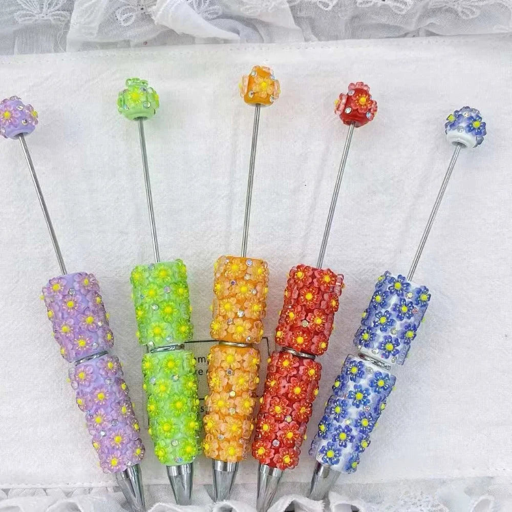 Five colorful daisy floral clay beadable pens in purple, green, orange, red, and blue with yellow rhinestone flower details and silver metal tips, arranged in a fan layout on a white fabric background.
