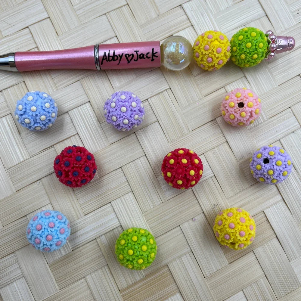 Colorful daisy flower ball resin fancy beads in blue, purple, red, green, yellow, and pink arranged on a woven surface next to a pink beadable pen with flower ball beads.