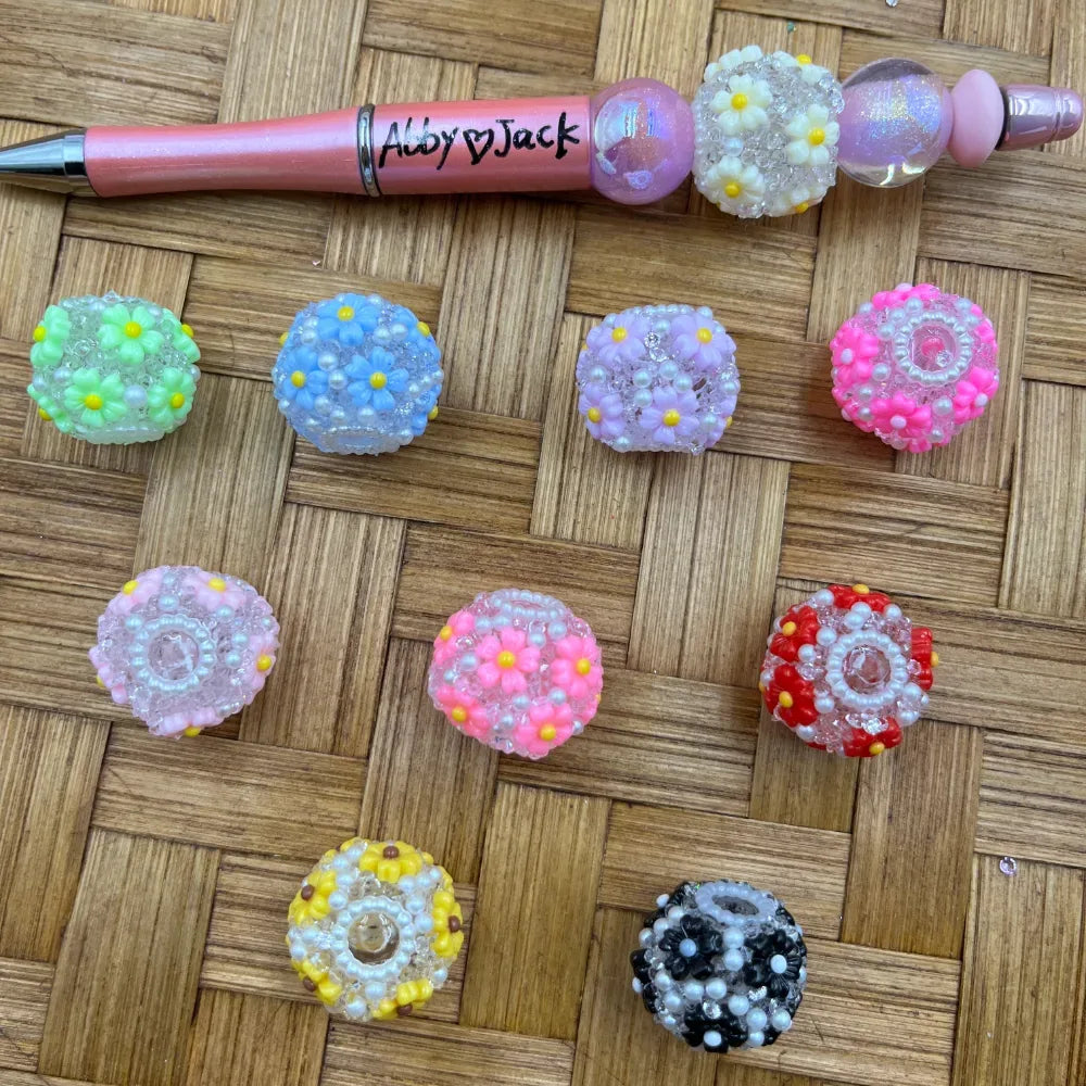 Assorted colorful daisy flower beaded ball beads in green, blue, lavender, pink, hot pink, red, yellow, and black displayed on a wicker surface next to a beadable pen labeled Abby and Jack.
