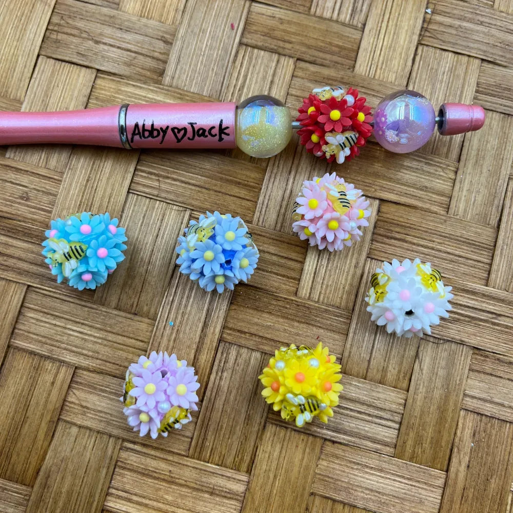 Assorted pastel daisy flower cluster fancy beads with miniature bumblebee accents in blue, pink, purple, yellow, white, and red, displayed on a wicker surface next to a pink beadable pen labeled Abby Jack.