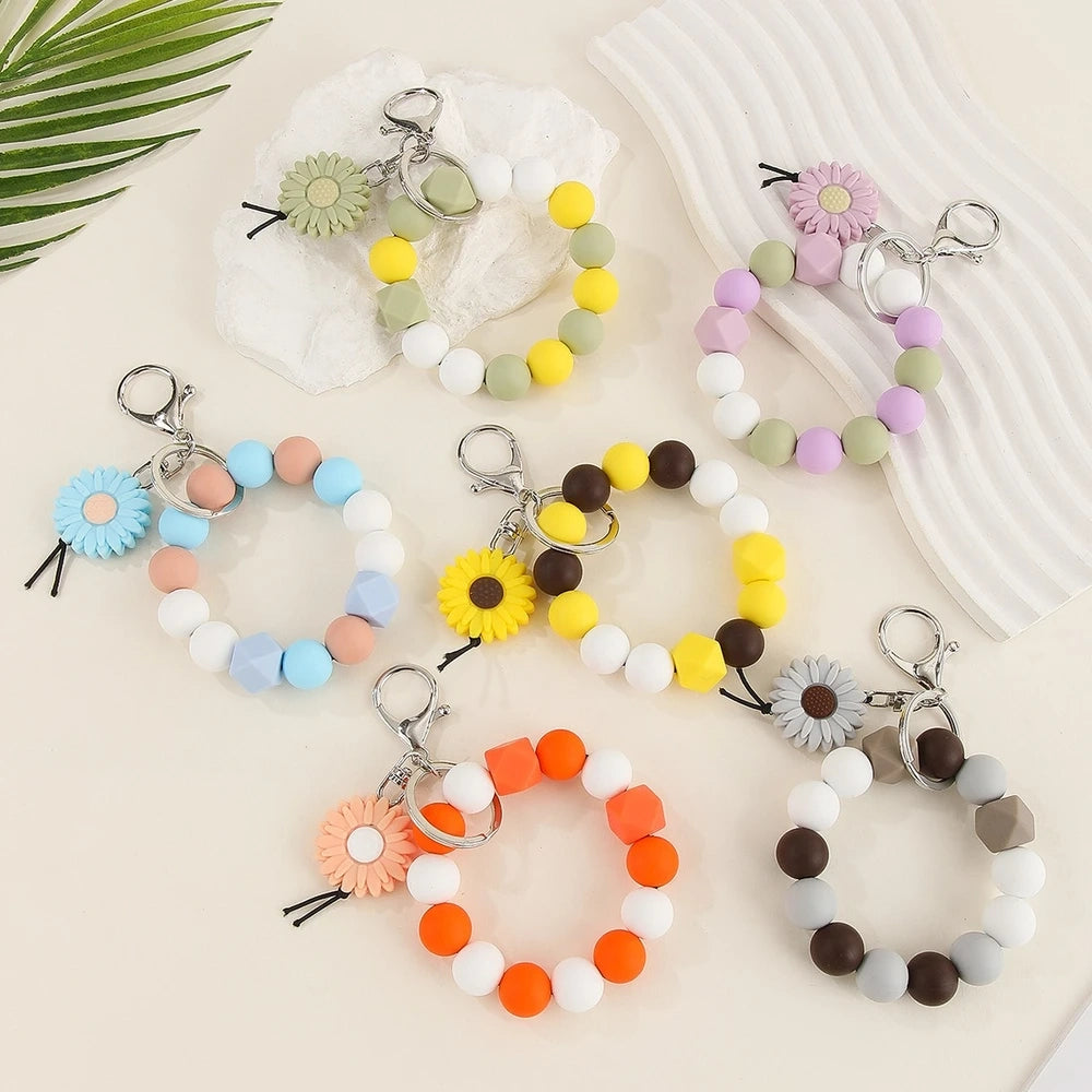 Seven colorful silicone bead keychain bracelet rings with daisy flower charms in various color combinations including orange, blue, yellow, green, purple, gray and brown on a white surface.