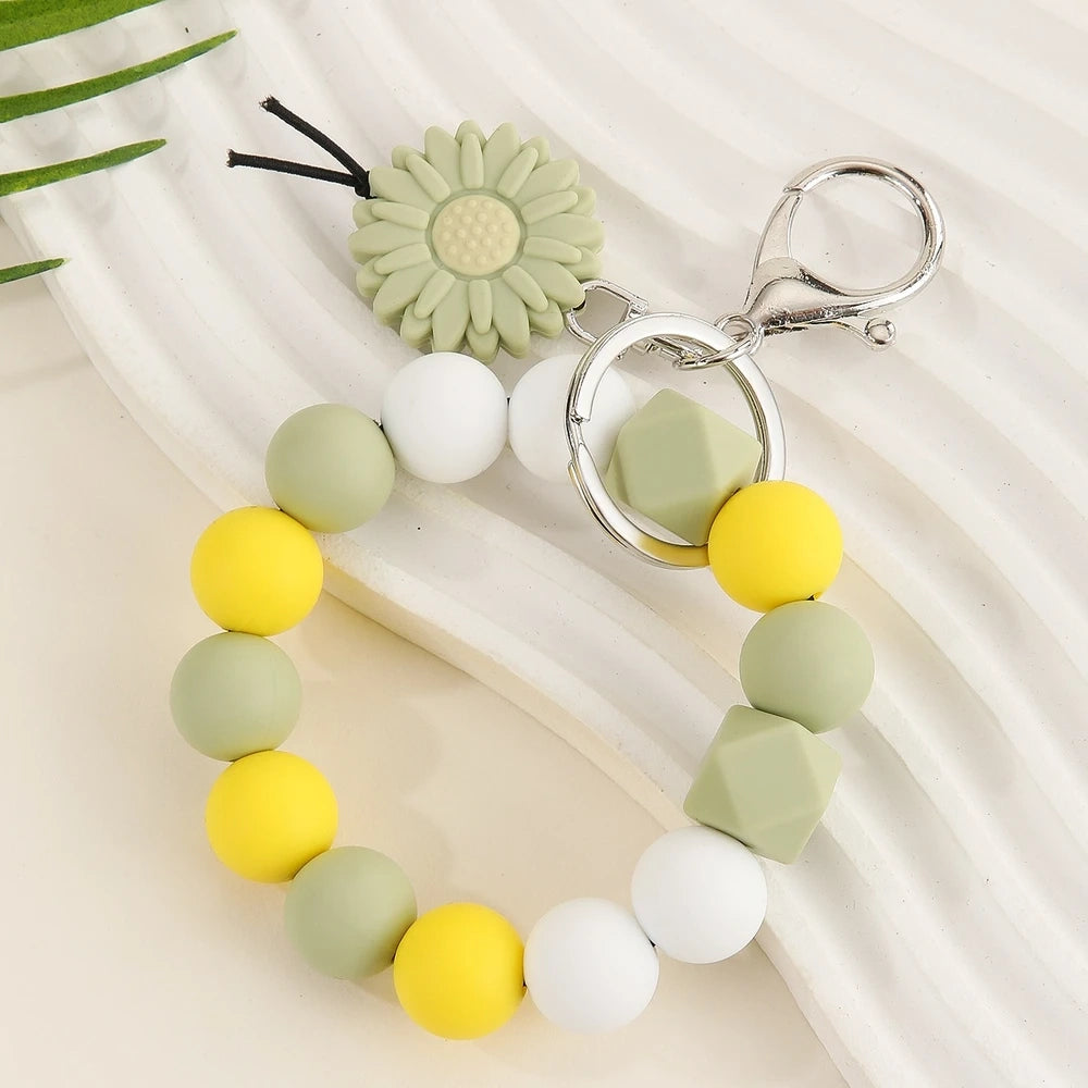 Seven colorful silicone bead keychain bracelet rings with daisy flower charms in various color combinations including orange, blue, yellow, green, purple, gray and brown on a white surface. - Green