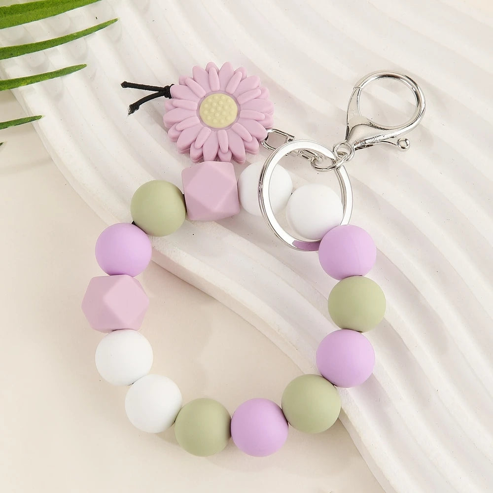 Seven colorful silicone bead keychain bracelet rings with daisy flower charms in various color combinations including orange, blue, yellow, green, purple, gray and brown on a white surface. - Purple