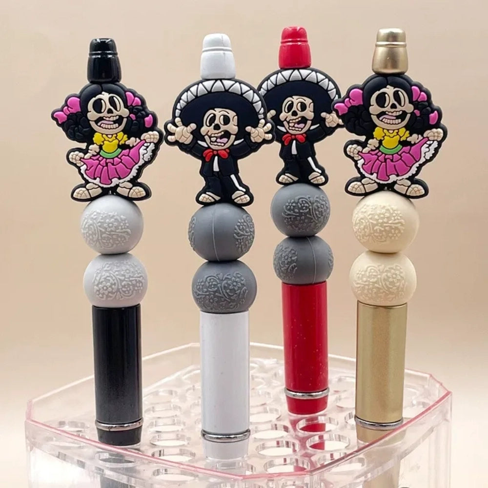 Four Day of the Dead skeleton silicone focal beads in black, gray, red, and gold colorways featuring Catrina and Mariachi skeleton characters, displayed in a clear acrylic pen holder. - Random Mixed