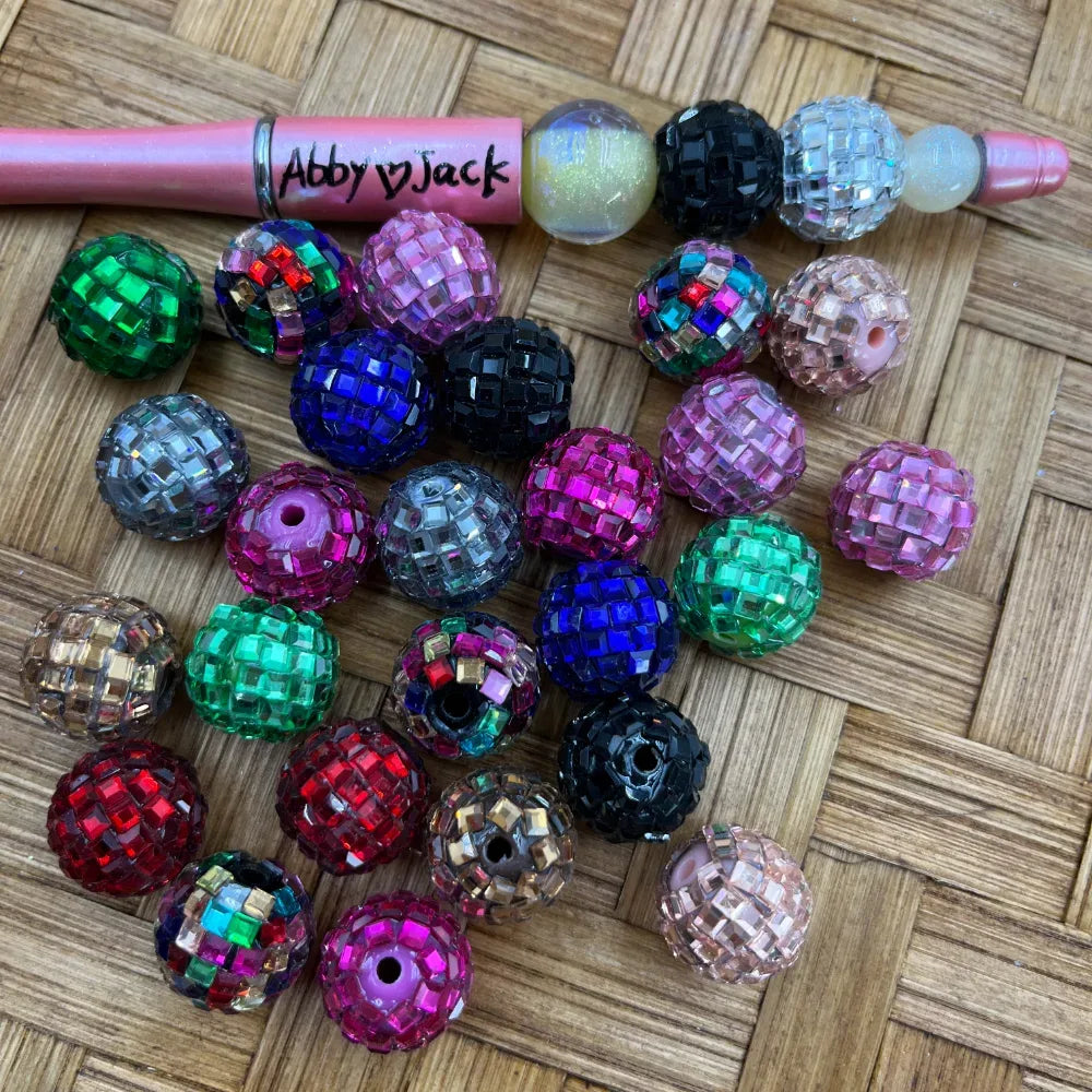 Assorted colorful disco ball rhinestone beads in red, green, blue, pink, black, silver, and champagne displayed on a woven surface next to a pink beadable pen branded Abby Jack.