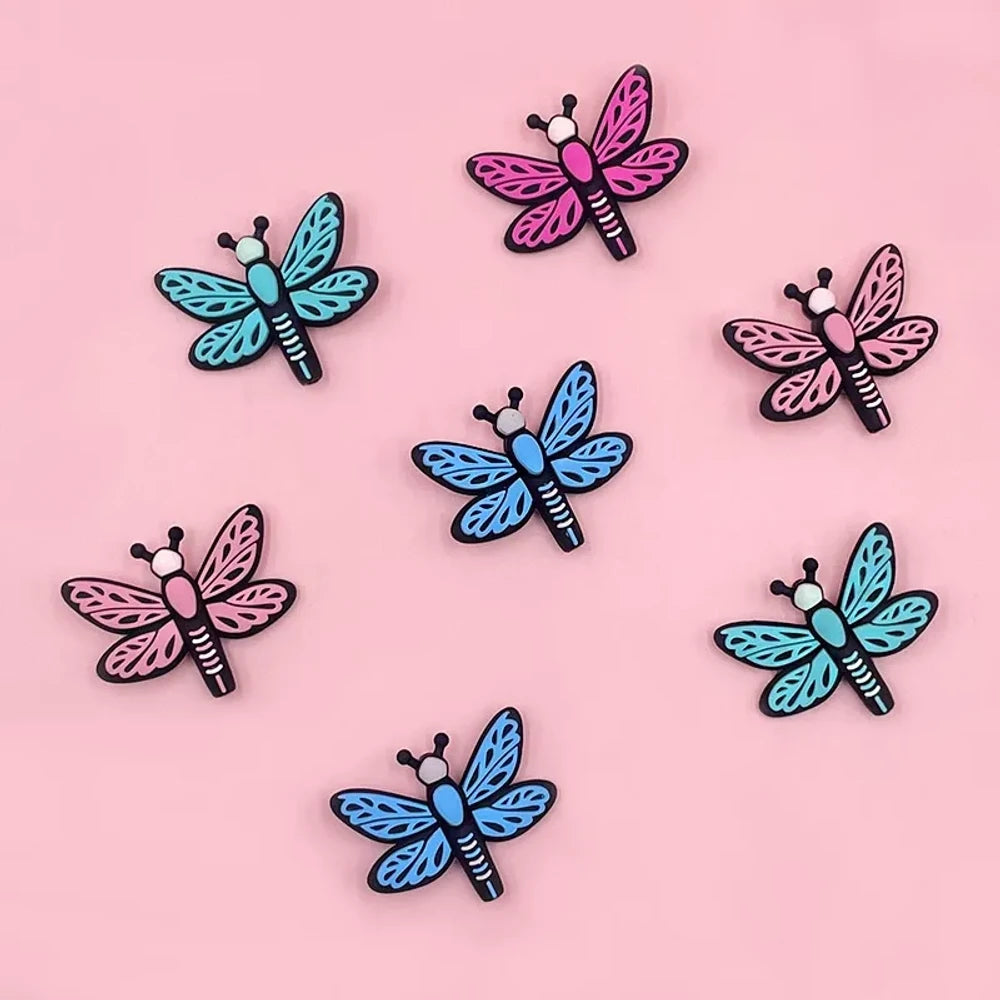 Seven colorful dragonfly silicone focal beads in pink, hot pink, light blue, and teal arranged on a pink background, featuring lace-pattern wings and black outlines.