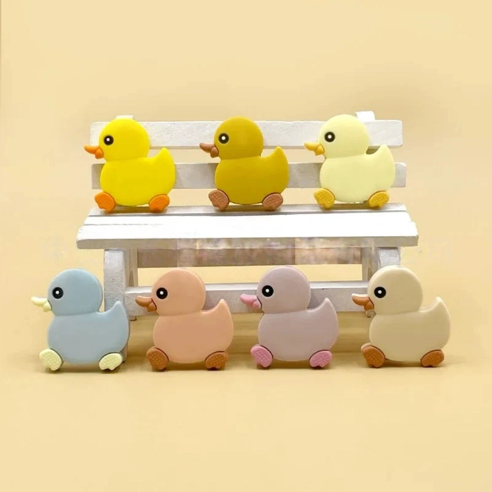 Seven cute duck silicone focal beads in yellow, mustard, cream, blue, peach, lavender, and beige colors arranged on a miniature white wooden bench, perfect for beadable pens and keychains.