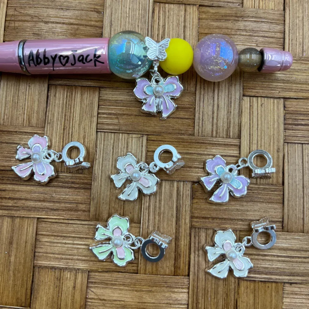 Silver-toned enamel bow flower charm bail beads in pastel pink, white, and iridescent finishes displayed on a wicker surface alongside a decorated beadable pen.