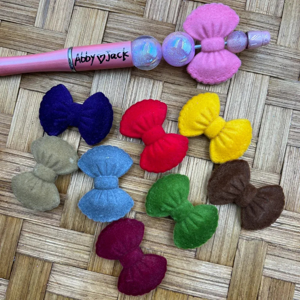Assorted color felt bow acrylic beads in pink, navy, red, yellow, tan, light blue, green, brown, and burgundy displayed on a woven surface next to a pink beadable pen labeled Abby Jack.