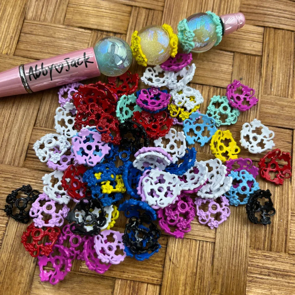 Pile of colorful filigree crown-shaped metal spacer beads in mixed colors including red, white, black, blue, pink, yellow, and teal, displayed next to a beadable pen on a wicker surface.