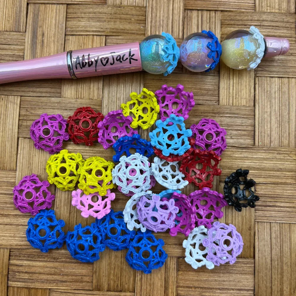 Assorted colorful filigree lace ball bead caps in pink, blue, yellow, red, white, purple, and black scattered on a wicker surface next to a pink beadable pen with glitter globe beads labeled Abby and Jack.