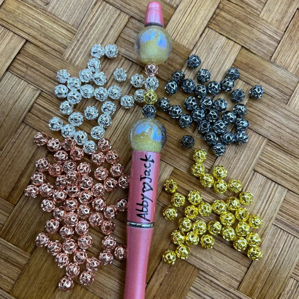 Four groups of filigree metal spacer beads in silver, black, gold, and rose gold arranged around a pink beadable pen on a woven bamboo surface, sold by DIY Mini Planet.