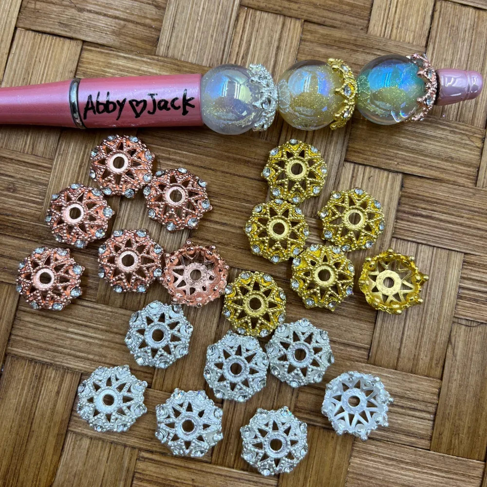 Assorted filigree rhinestone bead caps in rose gold, gold, and silver finishes displayed on woven surface next to a beadable pen, suitable for DIY jewelry making and beadable pens.
