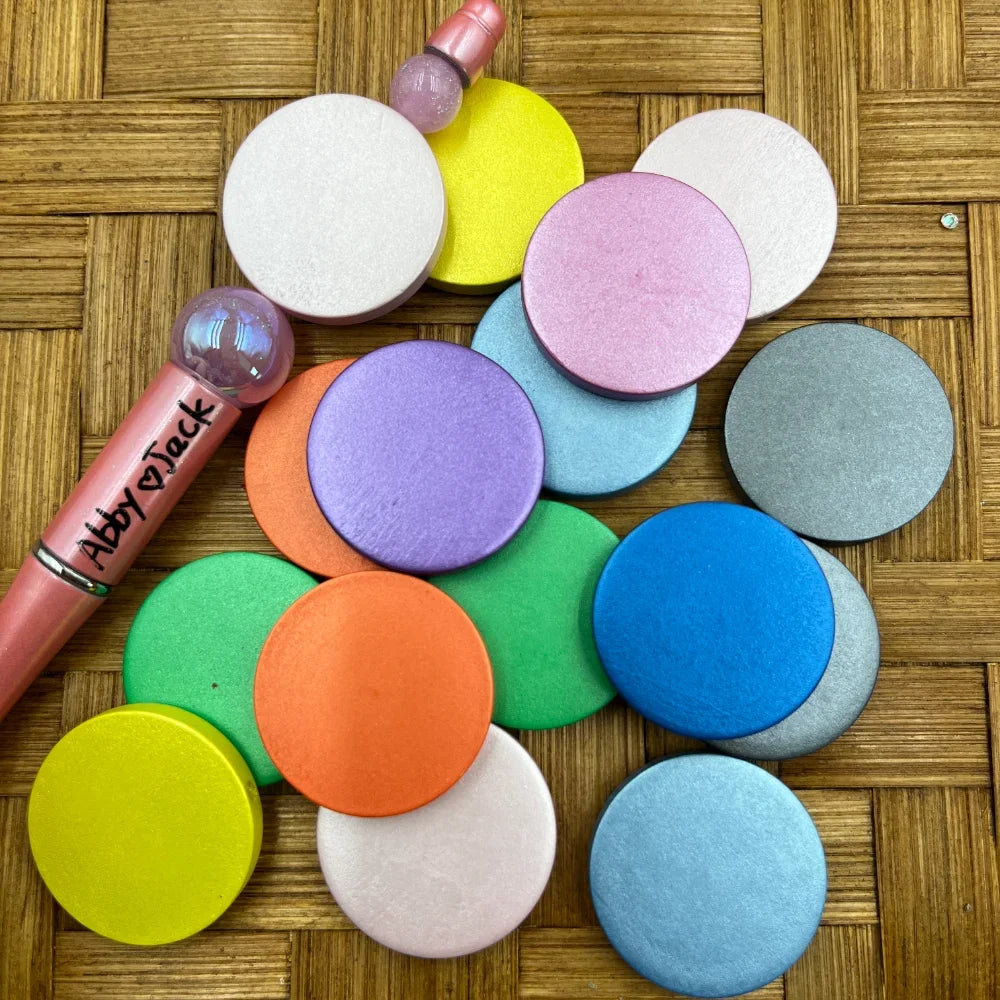 Assorted rainbow pastel flat round wood beads in colors including white, yellow, pink, purple, orange, green, blue, gray, and light blue displayed on a woven surface next to a branded beadable pen.