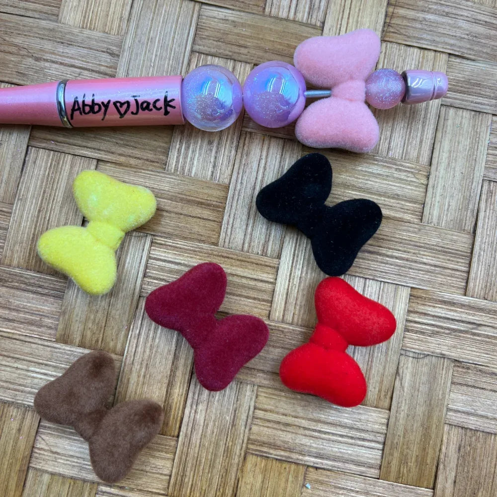 Assorted flocked velvet bow-shaped acrylic beads in pink, yellow, black, red, burgundy, and brown displayed next to a pink beadable pen on a wicker surface.