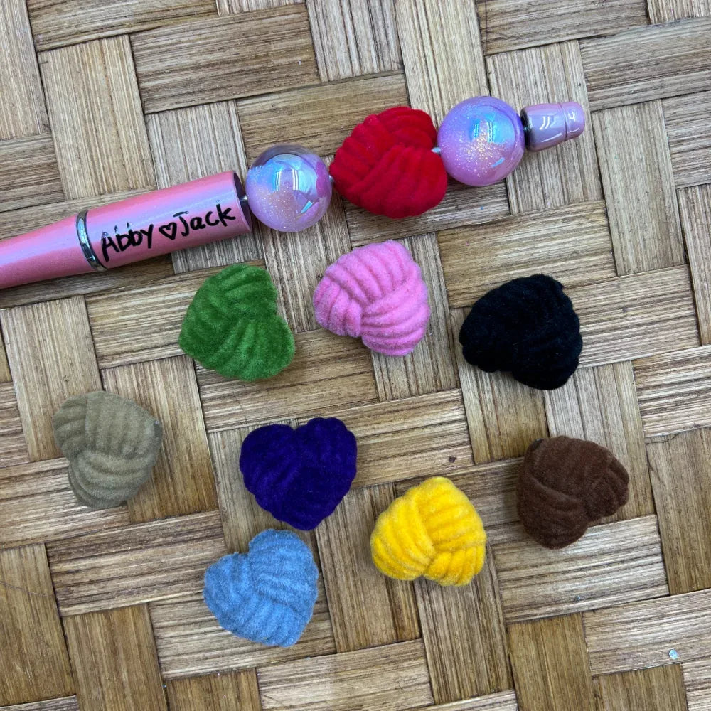 Assorted flocked velvet heart-shaped acrylic beads in red, pink, green, black, tan, navy, blue, yellow, and brown displayed on woven surface next to a pink beadable pen.