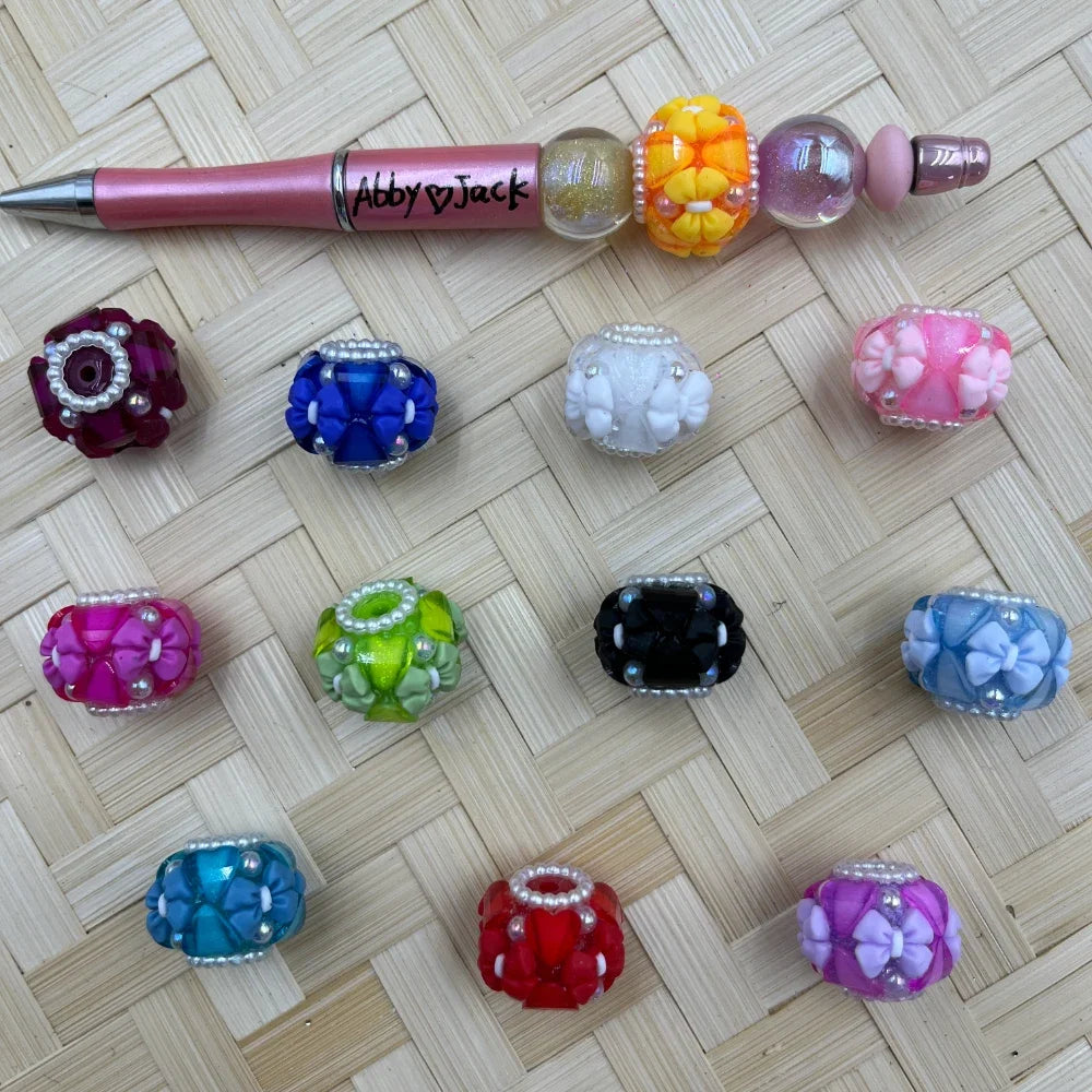 Assorted colorful floral bow fancy chunky beads with pearl accents in 12 colors including red, blue, pink, black, green, teal and purple, displayed on woven bamboo surface next to a pink beadable pen labeled Abby and Jack.