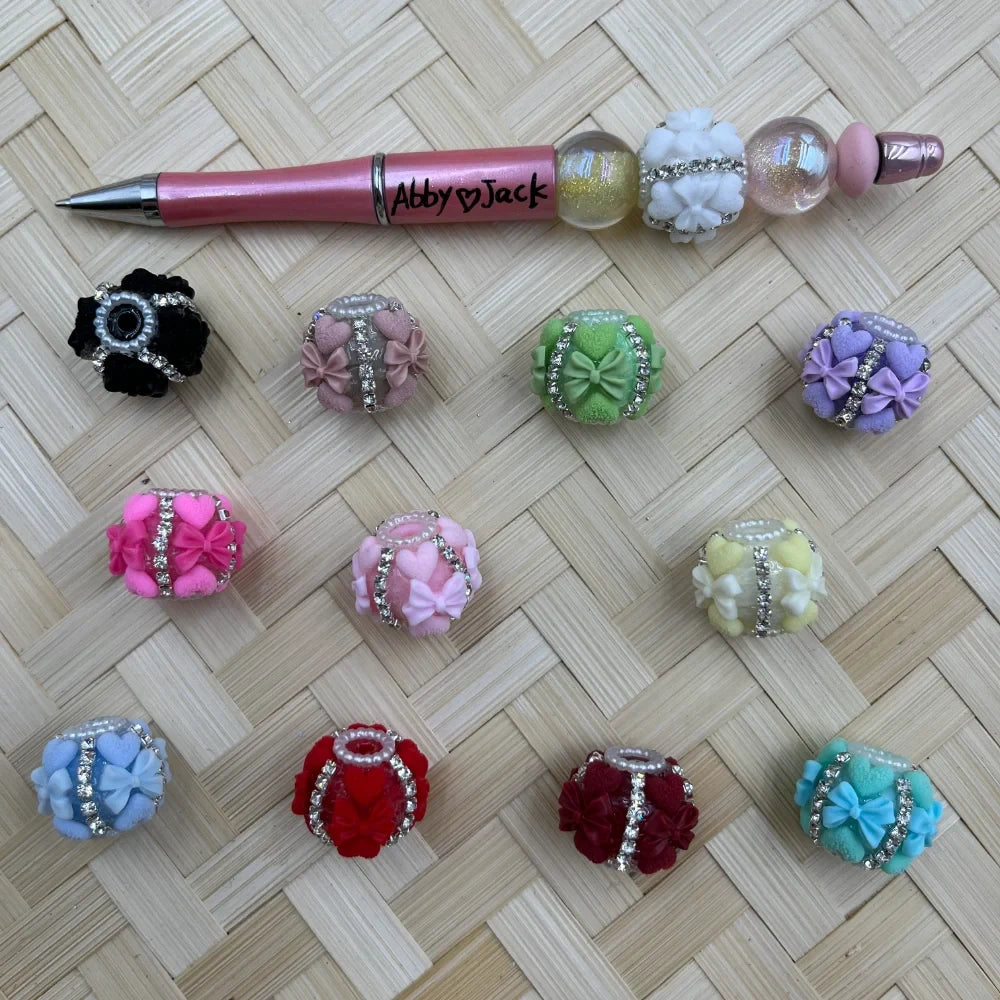 11 colorful fancy clay beads decorated with floral bows, rhinestone bands, and pearl accents in black, pink, green, purple, hot pink, light pink, yellow, blue, red, dark red, and teal, displayed on a woven bamboo surface next to a pink beadable pen labeled Abby Jack.