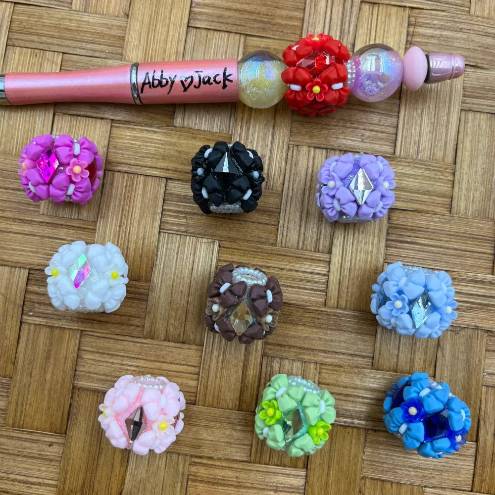 Assorted floral clay fancy beads with rhinestone centers in purple, black, lavender, white, brown, pink, green, blue colors displayed on woven surface alongside a decorated beadable pen labeled Abby and Jack.