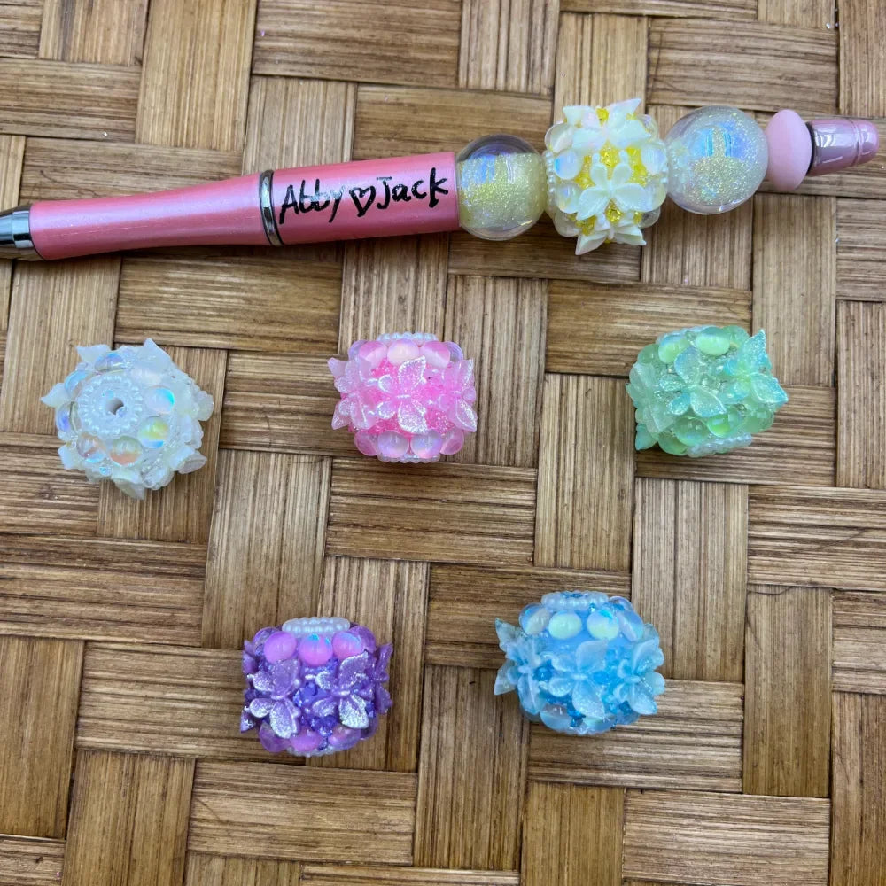 Assorted pastel floral cluster resin fancy beads in white, pink, green, purple, and blue displayed on a woven surface alongside a pink beadable pen labeled Abby and Jack.