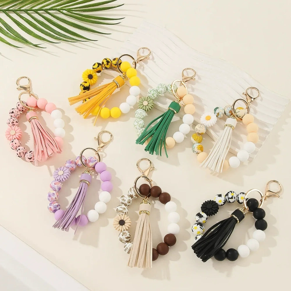Assorted floral and daisy printed silicone bead bracelet keychains in pink, yellow, green, purple, brown, white, and black colorways with tassel accents displayed on a cream surface.