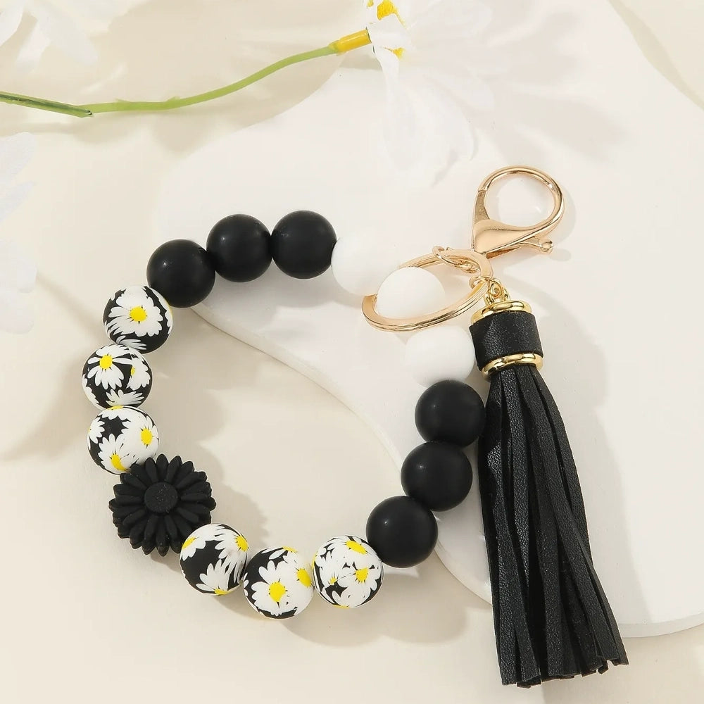 Assorted floral and daisy printed silicone bead bracelet keychains in pink, yellow, green, purple, brown, white, and black colorways with tassel accents displayed on a cream surface. - Black