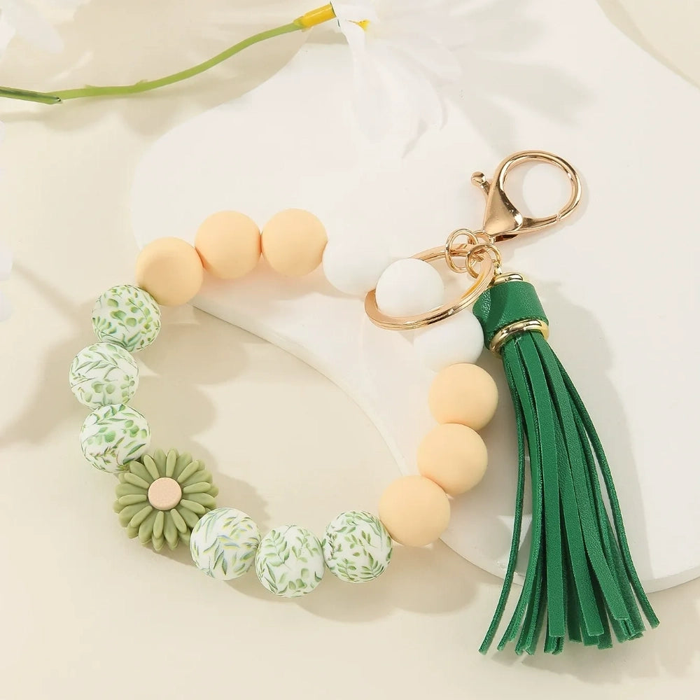 Assorted floral and daisy printed silicone bead bracelet keychains in pink, yellow, green, purple, brown, white, and black colorways with tassel accents displayed on a cream surface. - Green