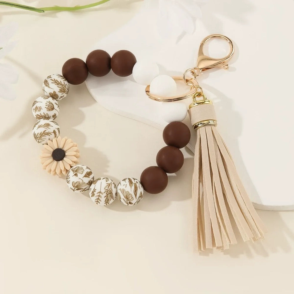 Assorted floral and daisy printed silicone bead bracelet keychains in pink, yellow, green, purple, brown, white, and black colorways with tassel accents displayed on a cream surface. - Khaki